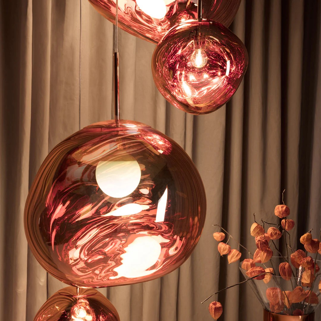 The Melt Trio Round Pendant System (Copy) showcases three glass pendant lights with a pink and gold marbled finish, suspended in front of beige curtains next to a vase of orange dried flowers.